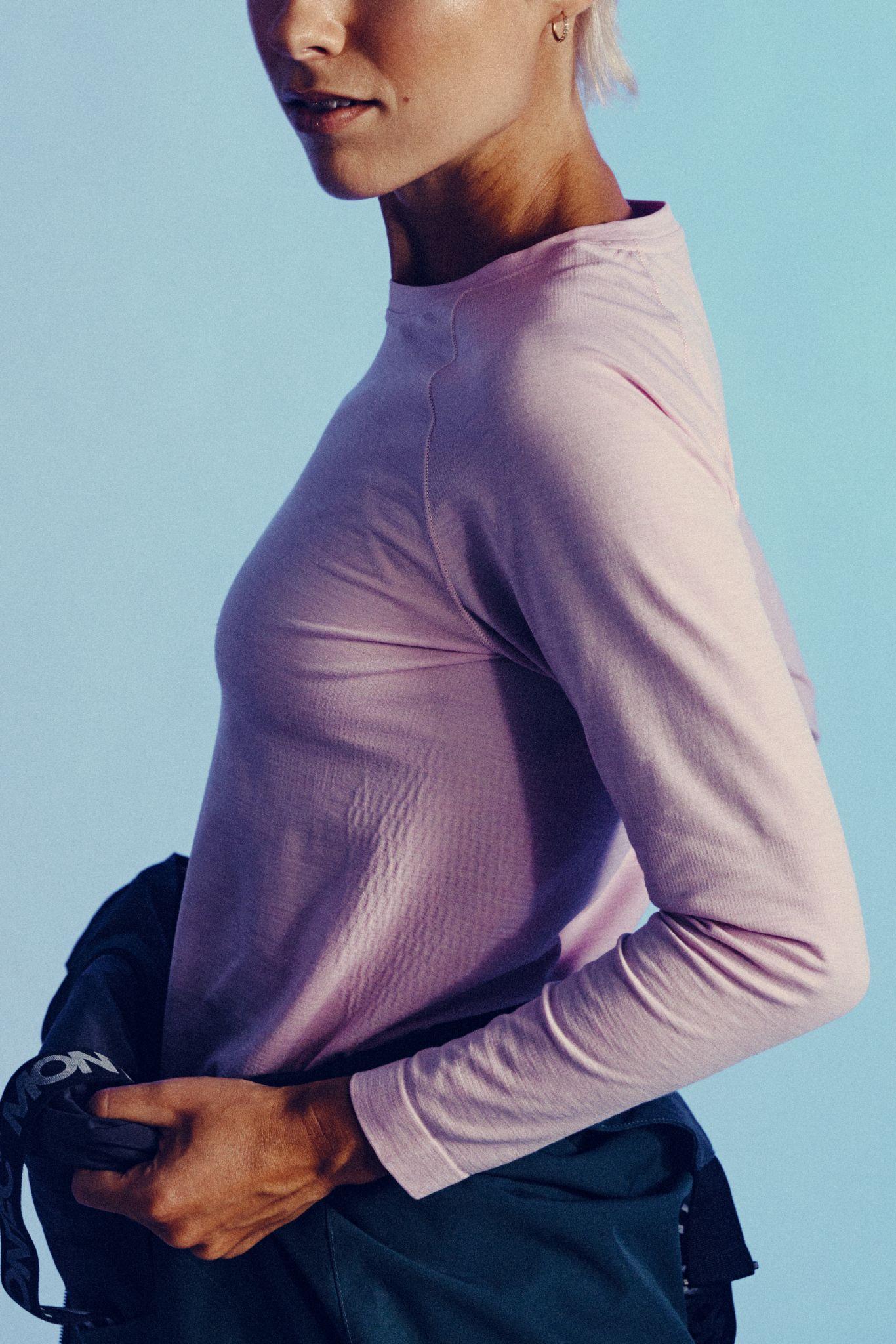 Woman with short hair in a light pink long-sleeve shirt holds a dark jacket against a light blue background. Only her upper body is visible in the image.