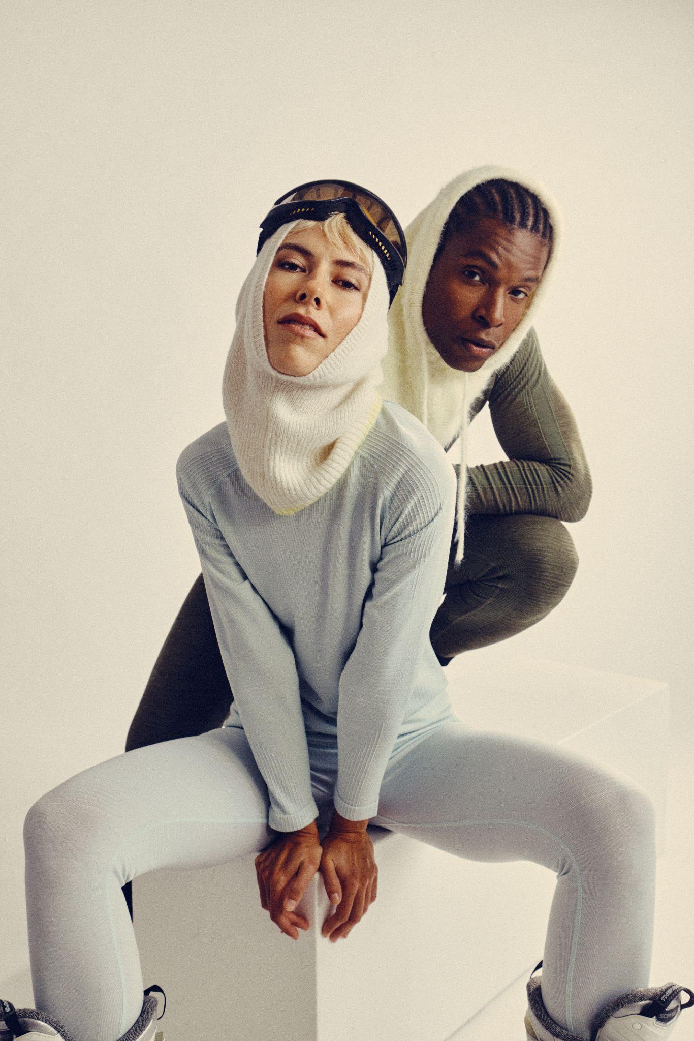 Two people in light-colored, fitted winter outfits with hoods and goggles pose stylishly against a plain background. One sits on a white block, while the other kneels behind, both looking confidently at the camera.