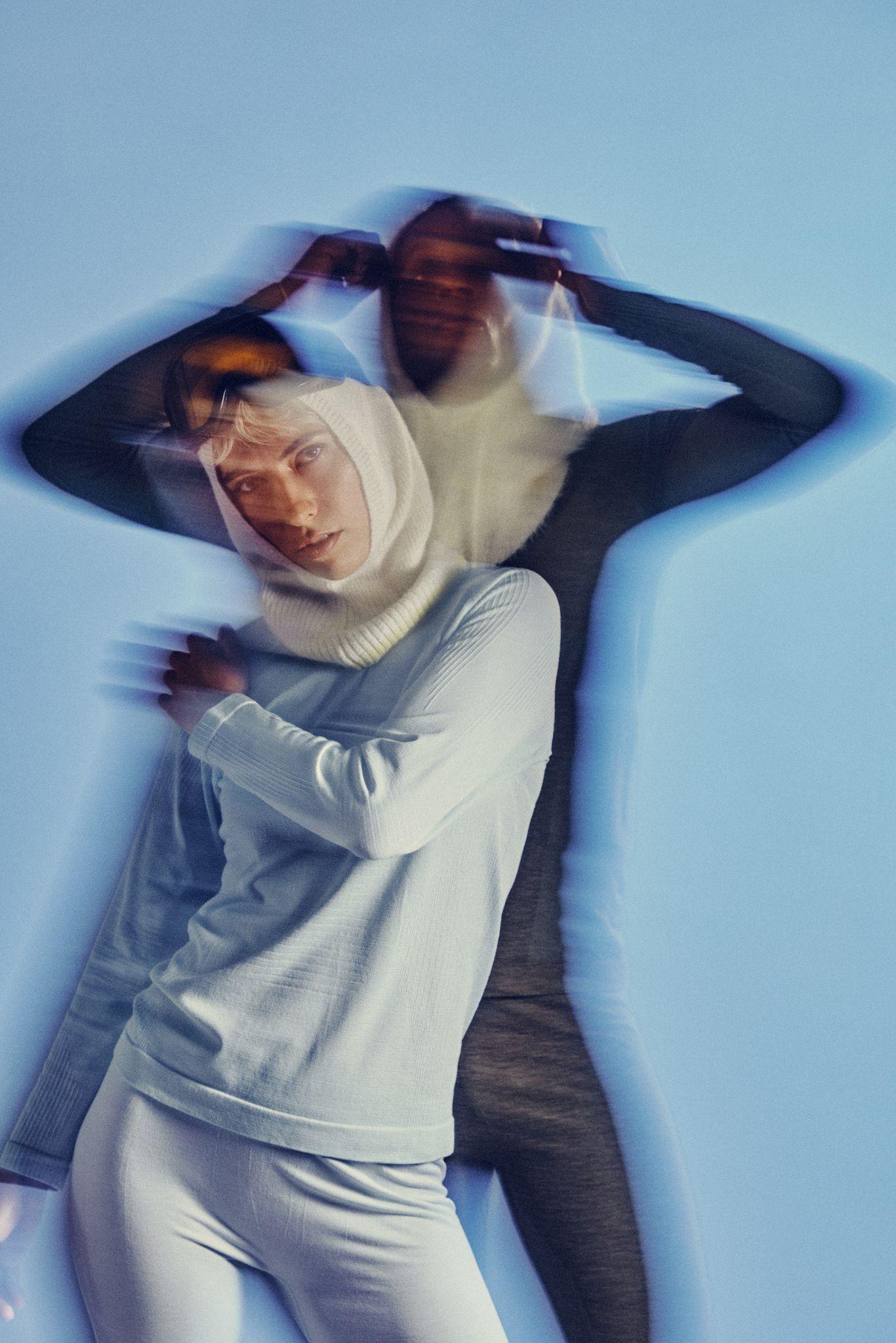 A person in light clothing and a white hood stands against a blue background, with a blurred, shadowy figure behind, creating a double exposure effect.
