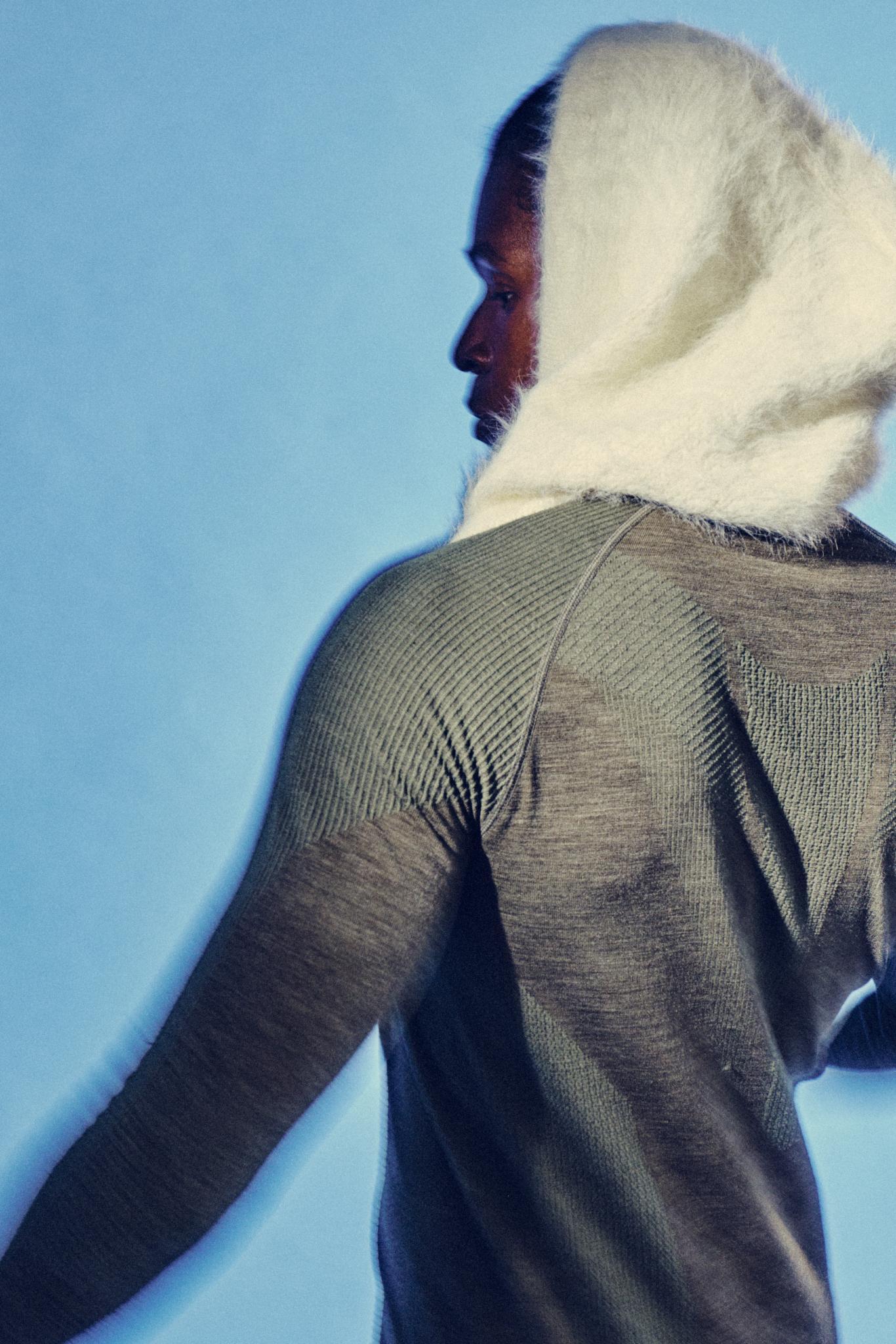 A person with a light knit hood over their head, wearing a fitted, textured gray sweater, is photographed from the back against a blue background.