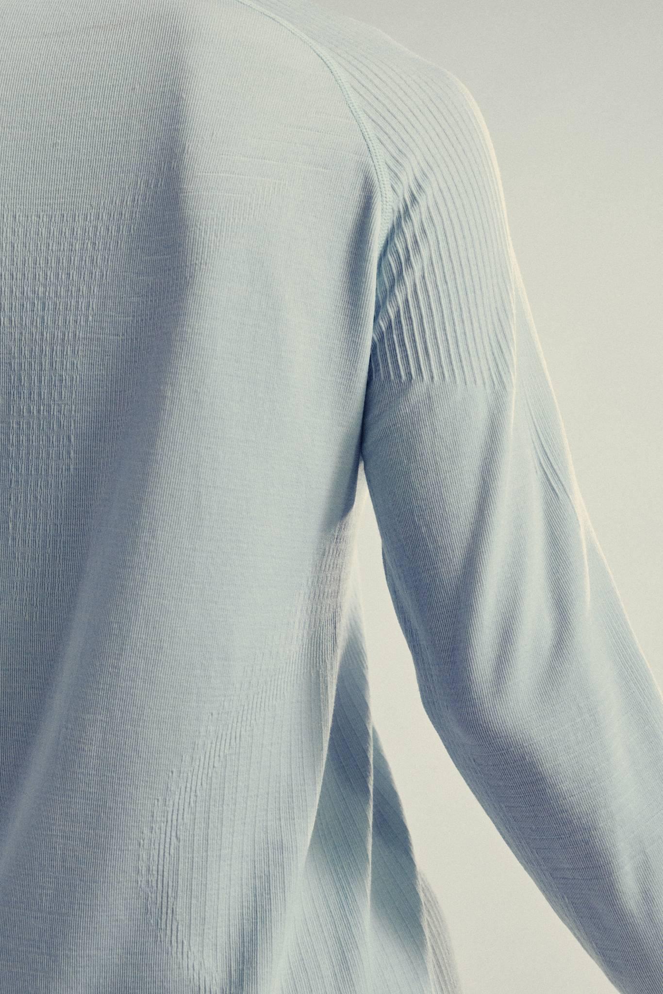 Close-up of a person wearing a pale blue, long-sleeved textured shirt, showing the upper back and part of the right arm with visible knit patterns. The background is softly lit and neutral.
