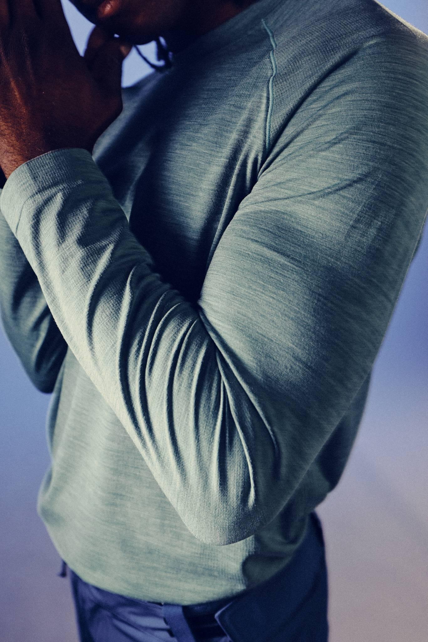 A person wearing a long-sleeve, light green shirt and dark pants stands with one hand near their face, partially visible, against a blurred background.