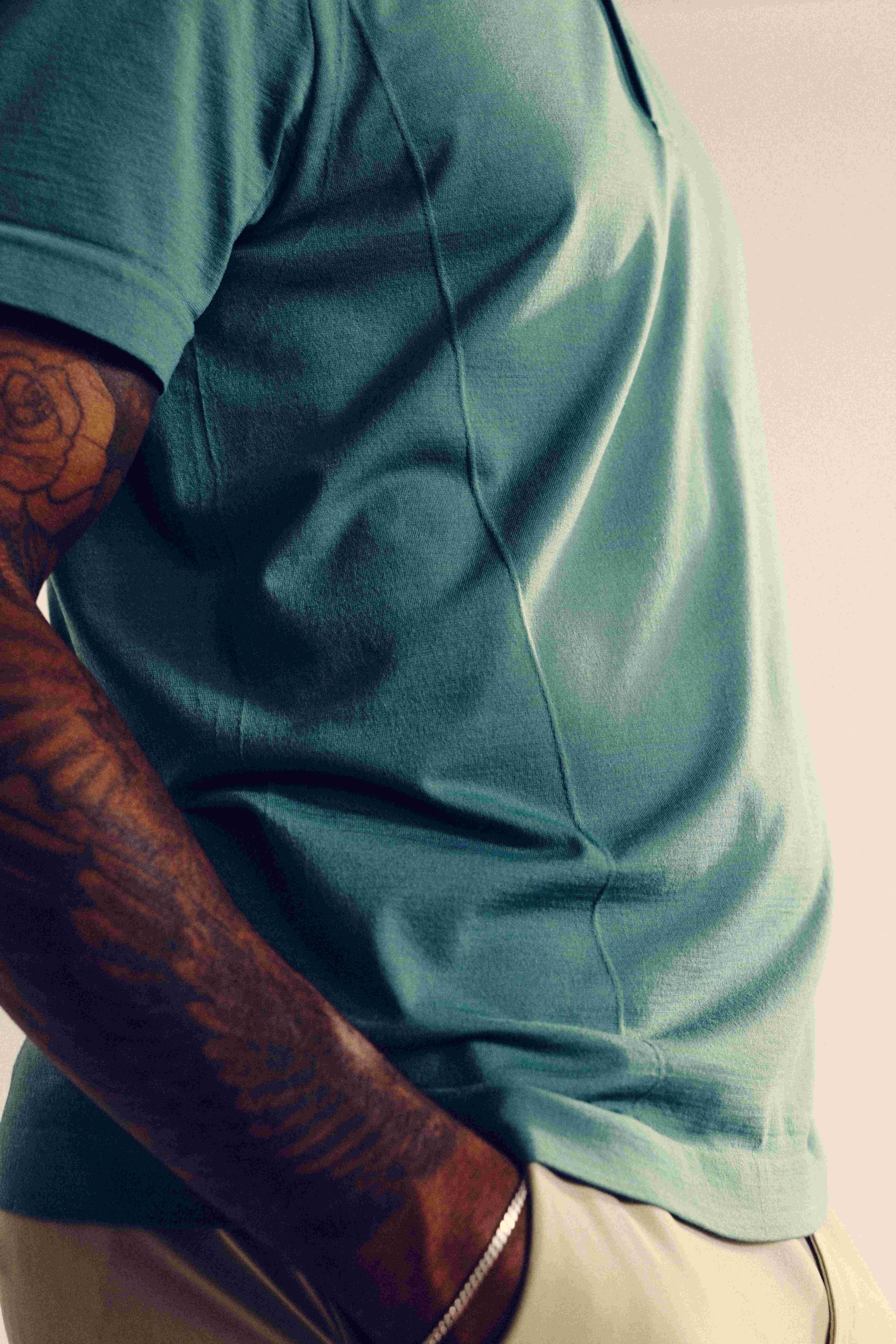 A person with tattooed arms wears a teal t-shirt and beige pants, standing with one hand in their pocket against a neutral background.