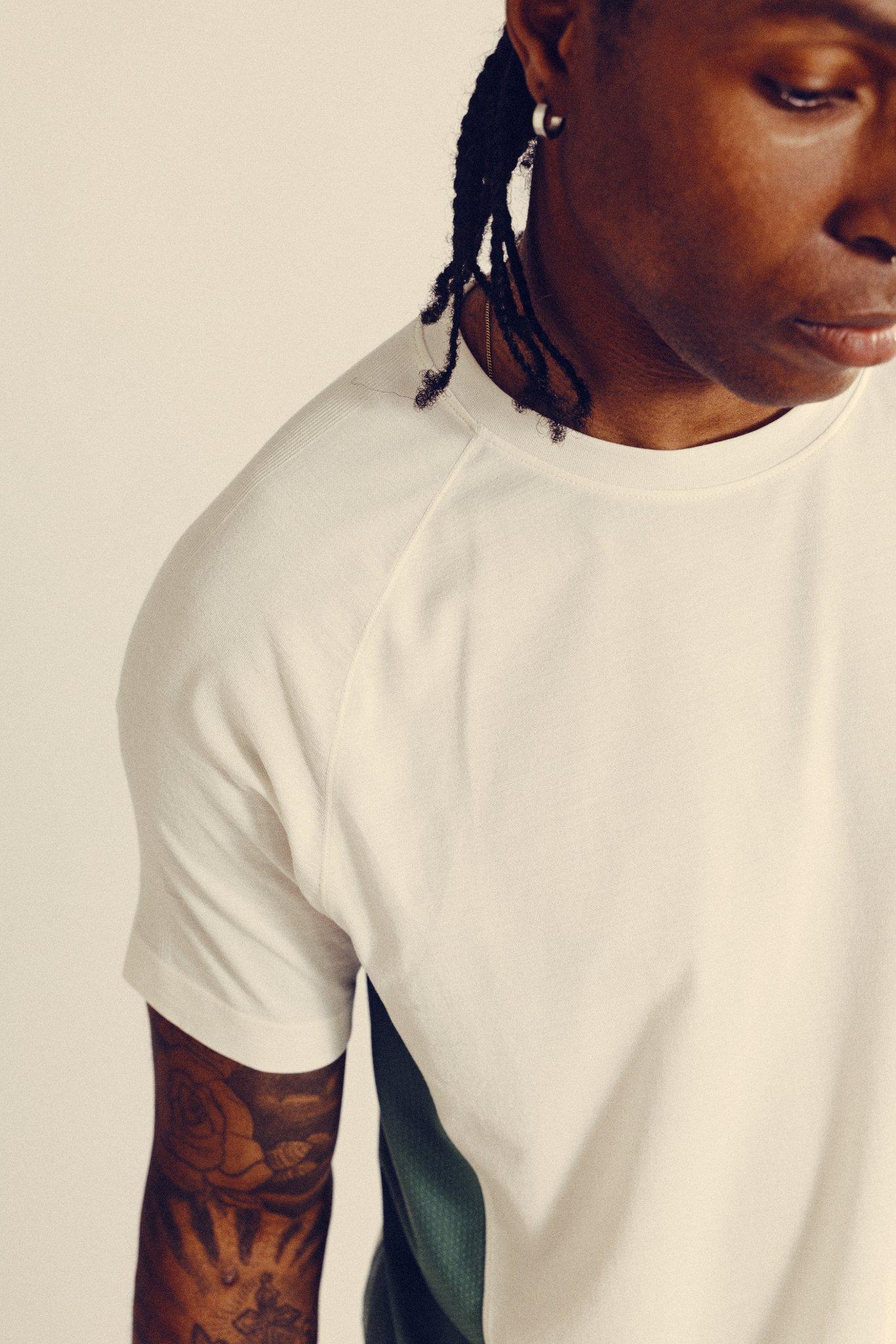 A person with braided hair and a tattooed arm wears a white short-sleeve shirt, looking downward. Only the upper body and part of the face are visible against a plain background.